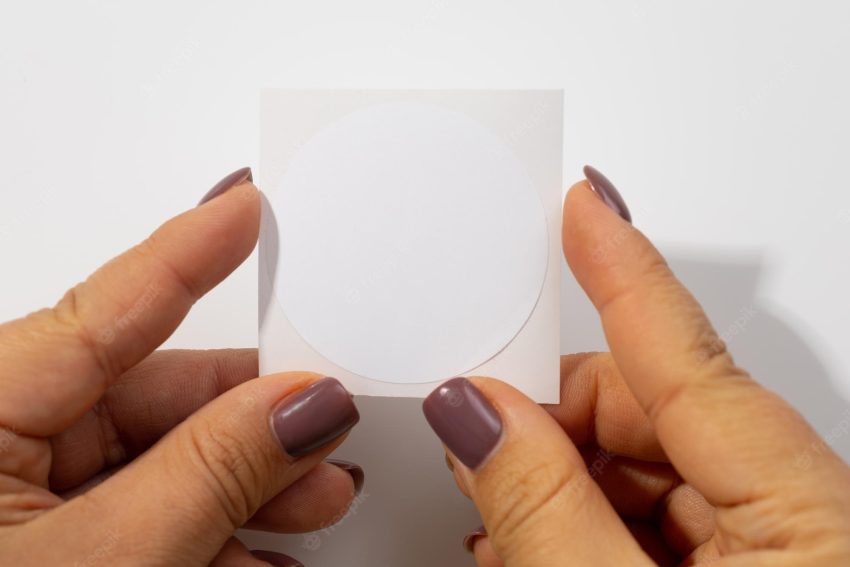 Circle sticky blank cardboard white label on white sheet of paper of square shape for clothes in woman hands on white background tag mock up copy space