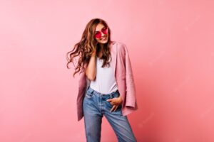 Excited white girl in bright stylish glasses posing on pink. dreamy curly woman playing with her ginger hair and laughing.