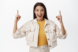 Portrait of cheerful brunette girl tell big news, woman pointing fingers up at sale banner, discount logo, smiling amazed, showing advertisement, white background. copy space