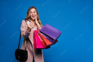 Young beautiful blonde woman in jacket on a blue wall with mobile phone doing online shopping.