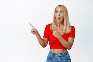 Amazed blond girl checking out big announcement in store pointing fingers and looking left at advertisement standing over white background