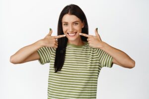 Attractive young woman pointing fingers at her smile showing white healthy teeth standing over white background in tshirt