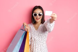 Beautiful asian woman in sunglasses going shopping holding bags and showing credit card standing ove