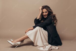 Cheerful model sitting on the floor, wearing modern oversize black jacket and creamy long dress, high heel shoes on her feet. curly hairstyle and makeup