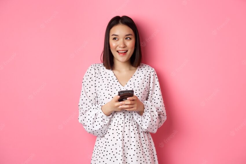 Cute asian woman thinking and smiling, looking left dreamy while messaging on smartphone, browing online stores, standing over pink background