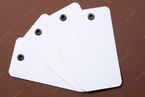 Four blank tags made of white cardboard of rectangle shape with tiny holes in upper part for clothes put in center on each other looking like fan on brown background tag mock up copy space