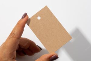 Hand with blank rectangular cardboard label tag for clothes with tiny hole in upper part in center with shadows falling on white background tag mock up copy space