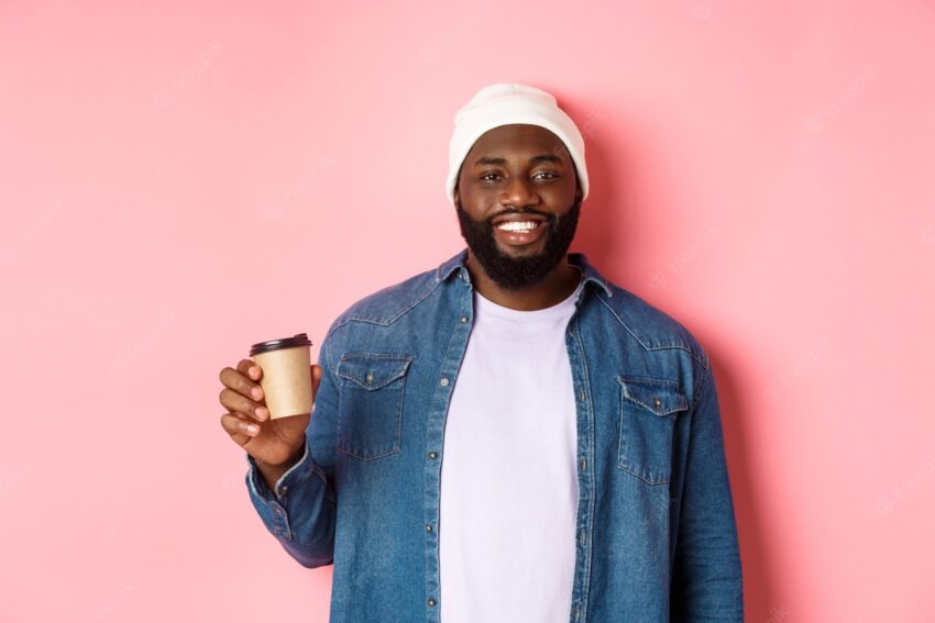 Handsome modern black man drinking takeaway coffee, smiling and looking satisfied at camera, standing over pink background