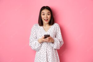 Online shopping. amazed asian woman looking at camera with happy smile, making purchase with smartphone, using mobile phone app, standing over pink background