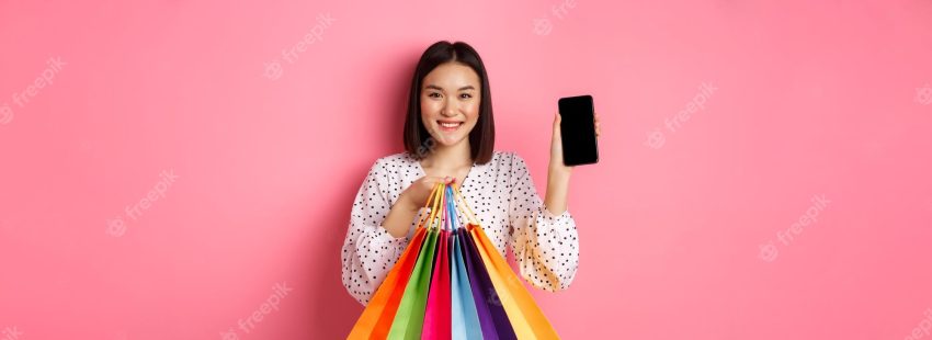 Attractive asian woman showing smartphone app and shopping bags buying online via application standi