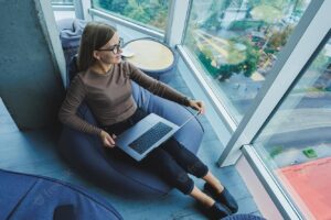 A beautiful female student works on a laptop while sitting on a bean bag chair in a modern creative office against the background of panoramic windows break time concept