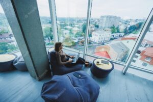 A beautiful female student works on a laptop while sitting on a bean bag chair in a modern creative office against the background of panoramic windows break time concept