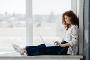 Beautiful positive woman using laptop sitting on the windowsill in city apartment young redhaired woman working at home freelance concept