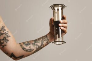Clear light gray aeropress with reflections held by a tattooed young man, close up portrait