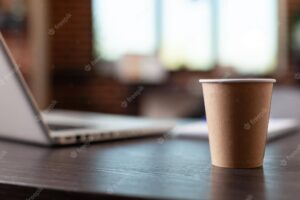 Close up of disposable coffee cup and laptop on office desk in startup space. hot beverage on table in empty business workplace. drink in plastic cup on furniture and computer.