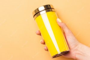 Female hand holding a yellow empty thermocup for drinks on a warm yellow background
