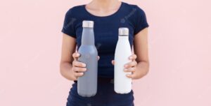 Two reusable steel thermo water bottles in female hands on the background of pastel pink color.