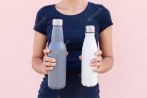 Two reusable steel thermo water bottles in female hands on the background of pastel pink color.