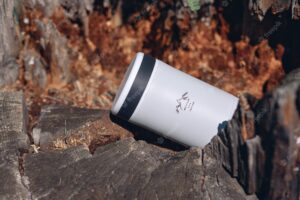 A white steel travel thermos with a print lies on a stump in close-up. a warm drink inside. a thing for tourism and hiking. camping concept.