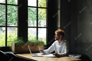 Young attractive smiling redhead bearded business man looking at window while sitting at workplace