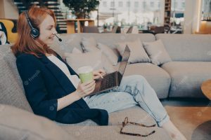 Young smiling female teacher working online via wireless headset and laptop sitting in cafe