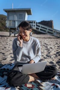 Young woman talking on smartphone while using her laptop at the beach