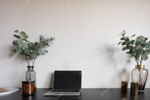 Bedroom working corner decorated with laptop white candles and artificial plant in glass vase on wood working table with beige painted wall  background