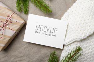 Christmas greeting card mockup with gift box, knitted sweater and fir tree branch on linen background