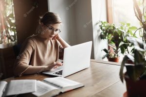 Cute european female coworker in glasses sitting near window in cafe leaning head on hand and reading articles in network via laptop searching information for work making notes in notebook