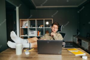 Freelancer lies in a chair at work at home with his legs raised on the table and uses a laptop