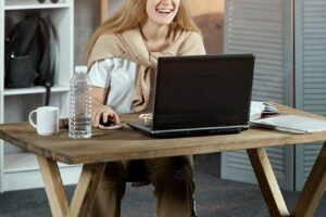 The girl is sitting at a table with a laptop and books on it, smiling carelessly