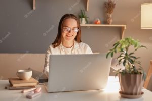 Horizontal shot of beautiful woman freelancer or student wearing white shirt and glasses sitting in home office in the evening working on laptop looking at screen
