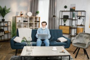 Man with grey beard mature male in casual clothes sitting on blue soft couch with laptop
