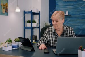 Mature woman using tablet and laptop in same time analysing financial graphs working from home sitting in workplace