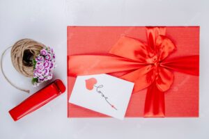 Top view of red gift box tied with bow and a small postcard a ball of rope with turkish carnation flower red stapler on white background
