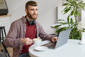Young attractive ginger bearded man working at a laptop, sitting in a cafe, wearing in basic clothes, broadly smiling and enjoy the work, looking away, drinking coffee and smiling.