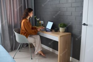 Young serious businesswoman in home wear sitting by desk in front of laptop