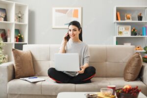 Young woman holding and used laptop speaks on phone sitting on sofa behind coffee table in living room
