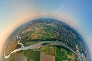 Aerial view from high altitude of little planet earth with small village houses and distant green cultivated agricultural fields with growing crops on bright summer evening