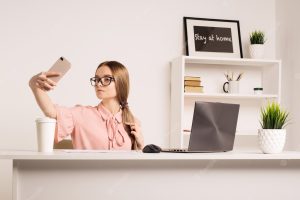 Portrait of a playful young girl taking selfie with mobile phone while sitting with laptop computer