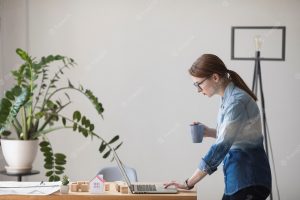 Side view of young woman holding coffee cup while working on laptop at workplace
