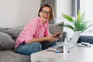 Smiling woman in pink shirt sitting relaxed on sofa at home at table working online on laptop from home