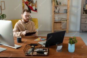 Young bearded creative male freelance designer sitting by table in front of laptop while using touchpad, stylus and color palette for drawing
